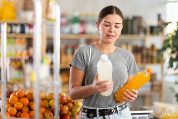 Fototapeta premium While shopping in grocery store, girl buyer choosing drink. Client look package of fruit drink, inspect product, reflect before buying. Customer examines contents of package, chooses beverage