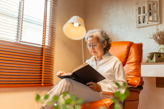 Wellness, mental healthcare in elderly people at home. Happy Asian senior mature woman relaxing on sofa in living room and reading a book. Retired woman enjoy leisure activity hobbies at nursing home.