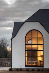 Obraz premium Modern architectural design features a striking white brick house with large arched windows against a cloudy sky, showcasing a beautifully illuminated interior at dusk