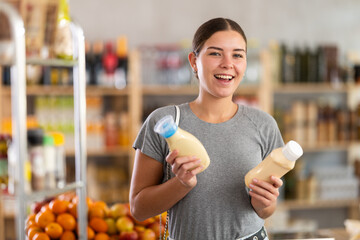 Fototapeta premium Joyful woman choosing packages of mayonnaise sauce in the supermarket. Buyer in the food section chooses two sauces, reading the composition and expiration date