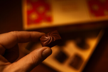 Closeup of a hand holding a chocolate praline with a box of chocolate pralines in the background. 