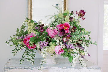 Floral arrangement in white textured compote with pink peonies, irises spirea, tulips, hydrangeas, lilacs on a table in front of a mirror