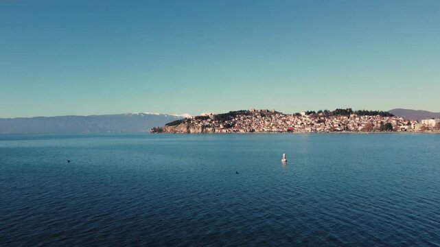 A lone seagull perched on a buoy in Lake Ohrid, North Macedonia. The picturesque town of Ohrid and snow-capped mountains create a stunning backdrop.