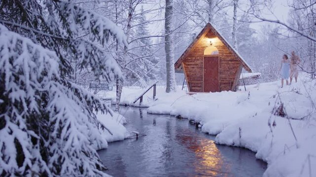 Couple in towels walk through snowy woods to wooden sauna next to stream, zoom