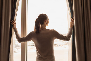 thoughtful woman looking out of her apartment window opening curtains 