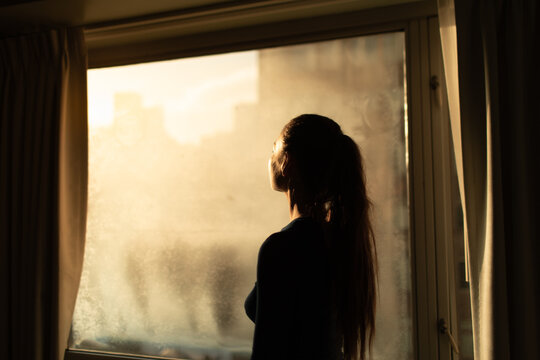 Woman in city apt looking out her window at the light of the sun from the darkness, sadness , hope, salvation, happiness concept 