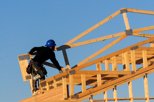Construction worker in a blue helmet assembling wooden roof trusses on a house frame under a clear sky. Represents home construction, framing, and structural building processes.