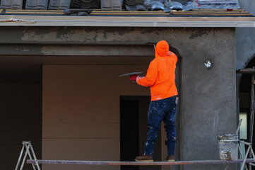 Fototapeta premium Construction worker in an orange hoodie plastering an exterior wall of a home under construction with a trowel while standing on scaffolding. Concept of construction and wall finishing in progress.