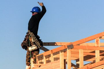 Construction worker in safety gear, including blue helmet and toolbelt, gesturing while framing wooden roof trusses under clear sky, representing homebuilding and residential construction industry.