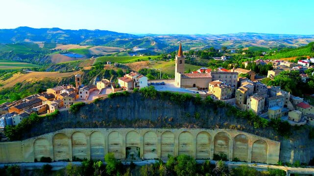 Picturesque aerial pullback shot from Castignano (AP), the town centre and its spired belltower, buildings perched on different levels above a steep hill supported by stunning arched walls underneath