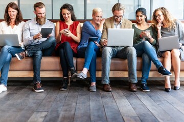 Group of diverse business startup people sitting on a bench using laptops and phones, engaged in technology and communication, in a casual office setting. Diverse team of people working on laptops.