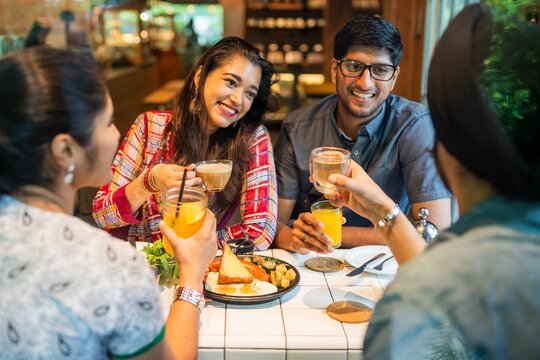 Group of friends enjoying drinks and food. Smiling and chatting, they raise glasses in a toast. Diverse group, relaxed atmosphere, cheerful gathering. Young Indian friends eating lunch at restaurant.