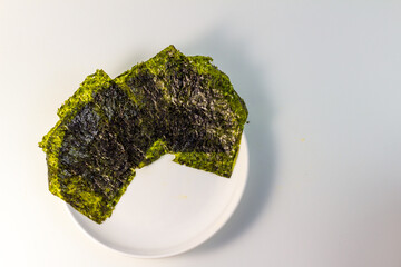 A close-up of a white plate with several sheets of dried nori seaweed. The nori sheets are a deep green color and have a slightly wrinkled texture. The plate is isolated on a white background.