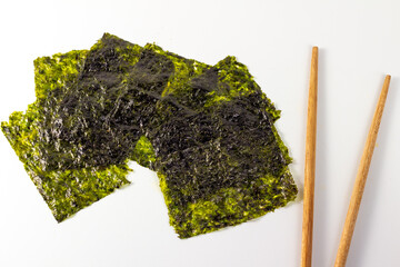 Several sheets of dried nori seaweed are scattered on a white surface. A pair of wooden chopsticks is placed to the side. The nori sheets are a deep green color and have a slightly wrinkled texture.