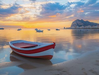 Photo of a small red and white boat on the beach at sunset in Alicante, Spain. The sky is reflected on the calm water with boats floating in the background