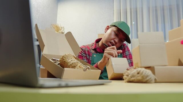 Young male business owner takes customer calls at home office. Managing shipment boxes while coordinating orders efficiently. home office, order fulfillment, entrepreneur work, shipping management
