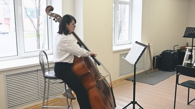 Double bass player rehearses near a large window, filling the room with deep, resonant tones. Her connection to the instrument reflects years of practice and dedication