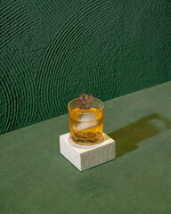 A Glass Of Cocktail Featuring A Round Ice Sphere, Topped With A Decorative Dark Brown Garnish, Elegantly Presented On A Minimalist White Block.