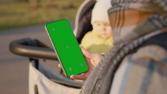 Mother holding smartphone with green screen standing next to baby stroller in park, showcasing mobile technology and parenting lifestyle
