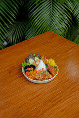 Fried Chicken Served with Rice, Crispy Skin, Egg, Tofu, Tempe, Crispy Sprinkles, Fresh Salad, and Spicy Sambal on a White Plate, Placed on a Wooden Table.