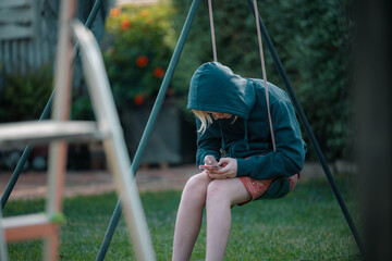 Pre-teen boy wearing hoodie relaxing on backyard swing looking at phone