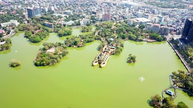 Aerial view of Kandawgyi Lake and Karaweik Palace in Yangon, Myanmar, showcasing the vibrant green water, lush greenery of the park, and the bustling cityscape in the background on a sunny day