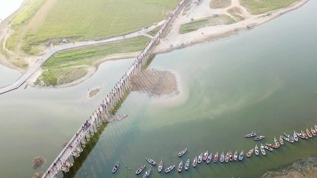 Aerial view of U Bein Bridge, a long teak footbridge spanning Taungthaman Lake in Amarapura, near Mandalay, Myanmar, with several boats waiting nearby and green fields in the distance