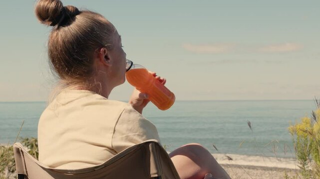 Woman drinks orange juice from bottle while relaxing on seashore sitting in tourist chair.