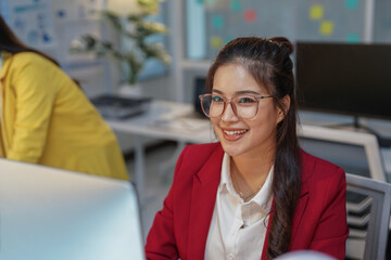 Young asian businesswoman wearing glasses and red jacket smiling and working on computer in modern office, with colleague in the background