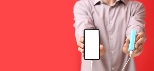 Young man holding mobile phone and power bank on red background with space for text, closeup