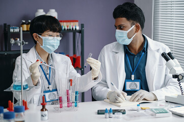 Two scientists conducting research in laboratory, wearing masks and white lab coats, examining...