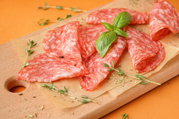 Wooden board with slices of tasty salami on orange background
