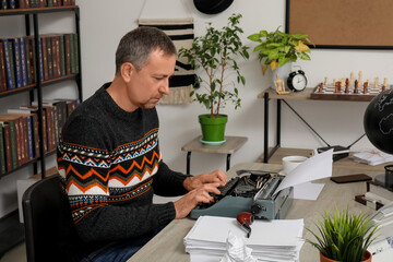 Thoughtful mature man typing on vintage typewriter and sitting at table in office