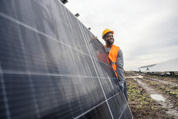 Smiling hardworking black worker carrying solar panel at solar farm.