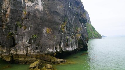 A close-up view of Ha Long Bay's weathered limestone karst reveals intricate textures and sparse vegetation clinging to its sheer cliffs, while the jade-green waters lap gently at its eroded base
