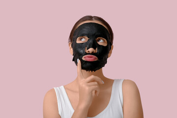 Thoughtful young woman with activated charcoal mask on her face against pink background