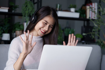 Asian woman sits on sofa wearing headphone using laptop video call with her friends in living room