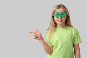 Cute little girl in decorative glasses in shape of clover pointing at something on grey background....