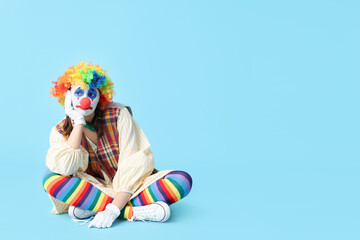 Young woman dressed as sad clown sitting on blue background. April Fool's day celebration