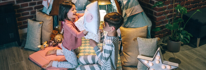 Banner of happy siblings having fun in pillow fight on night party at living room. Cheerful boy and girl with pajama laughing and playing in cushion battle at home. Domestic family life concept