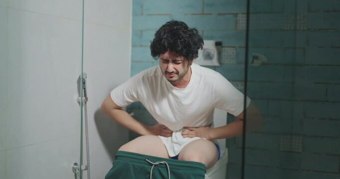 Indian asian young adult teen age gen z male boy guy alone upset tired sad man sitting on toilet bowl hold painful belly health issue feel unwell hurt chronic ache suffer facing sick pain indoor home