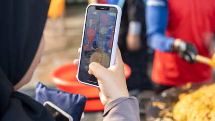 A close-up of a person taking a photo of street food with a smartphone, capturing a vendor preparing stir-fried noodles in an outdoor market setting.
