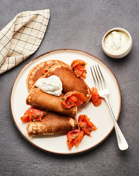 Savory pancakes are elegantly presented on a white plate with smoked salmon and a dollop of cream on top, creating an appetizing dish. A fork and checkered cloth complement the setting