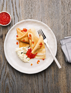 A plate holds folded crepes garnished with vibrant caviar and accompanied by a dollop of fresh cheese. A stylish fork rests beside the dish on a wooden surface