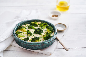 Broccoli casserole is freshly baked in a blue-green ceramic dish on a wooden table. Olive oil and spices accompany this comforting and healthy meal, perfect for family dinners