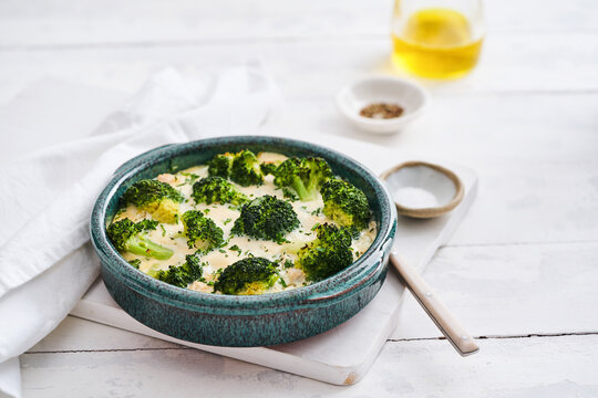 Broccoli casserole is freshly baked in a blue-green ceramic dish on a wooden table. Olive oil and spices accompany this comforting and healthy meal, perfect for family dinners