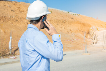 A construction worker wearing a safety helmet is making a phone call at a busy construction site under clear blue skies