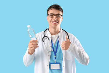 Young male doctor with bottle of water showing thumb-up on blue background