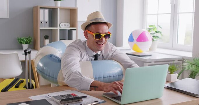 Happy businessman, entrepreneur or corporate employee in sunhat, sunglasses and swim ring sitting at office work desk, getting ready for summer vacation, typing on laptop, booking holiday package tour