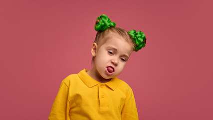 Preschool girl in yellow shirt with green braided buns hairstyle, sticking out tongue in playful mood against pink studio background. Concept of childhood, emotions, lifestyle